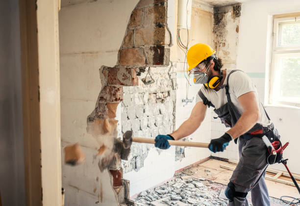 Строительный рабочий в защитном снаряжении сносит кувалдой кирпичную стену внутри комнаты.